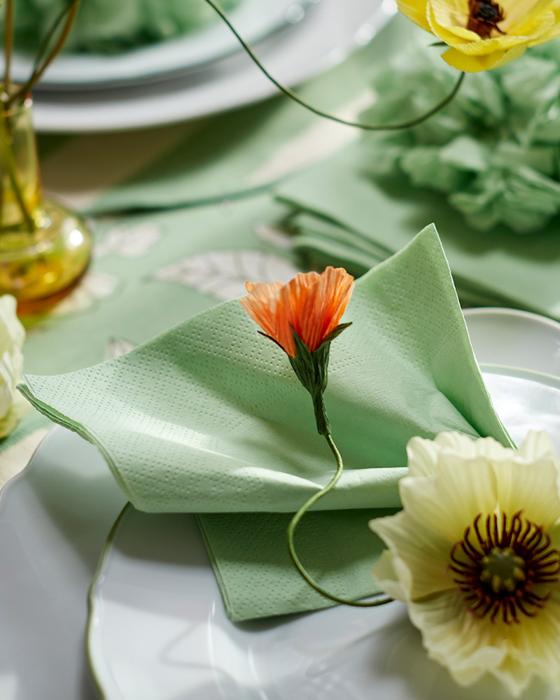 Una servilleta de papel verde FANTASTISK, sobre un plato y un plato de acompañamiento UPPLAGA, decorada con una rama artificial de magnolia SMYCKA.