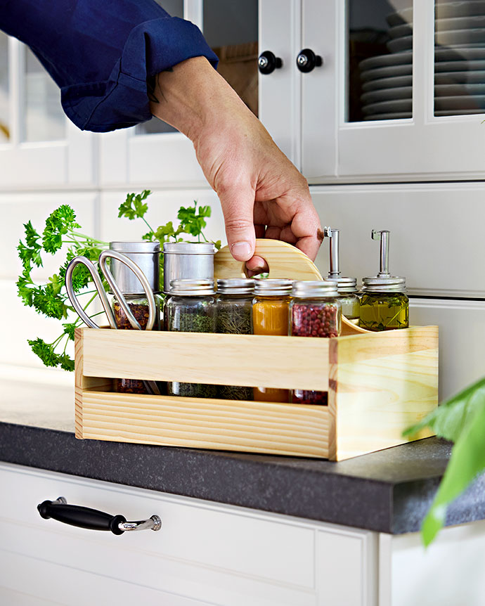 Hand lifting a wooden spice caddy with jars of spices, oil, and fresh parsley on a kitchen countertop.
