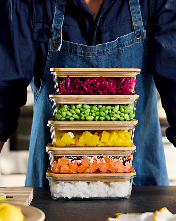 Stack of five glass and bamboo containers filled with rice, salmon, mango, edamame, and purple cabbage. Chef in blue apron.