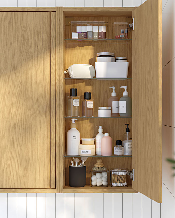 Primer plano de un armario de baño de madera abierto, mostrando organizadores transparentes y estantes de vidrio llenos de artículos de tocador.