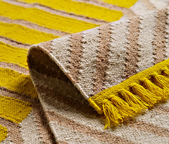 Detalle en primer plano de una alfombra de tejido plano con rayas amarillas y beige y acabado de flecos.