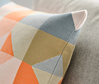 Detail of a cushion with a geometric pattern in orange, blue, and grey tones on a neutral sofa.