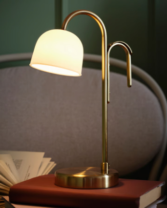 An opal white TVÅMASTAD LED portable lamp is elegantly placed on a stack of books on a cosy armchair.