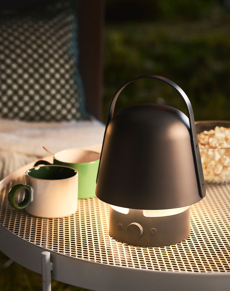 Un jardín cuenta con una lámpara con altavoz Bluetooth VAPPEBY azul de diseño redondeado, colocada sobre una mesa con bebidas.