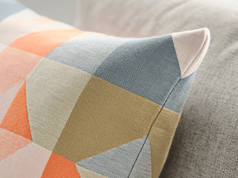 Primer plano de un cojín con estampado geométrico en tonos naranja, azul y mostaza sobre un tejido gris con textura.