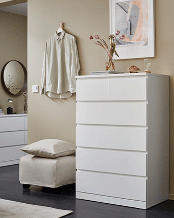 Cómoda blanca de 6 cajones en una estancia beige, decorada con un jarrón de flores y una camisa colgada en la pared.