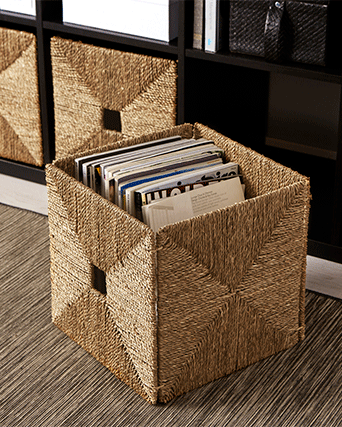 Cesta de fibras naturales tejida a mano con revistas, situada sobre una alfombra de rayas junto a una estantería oscura.