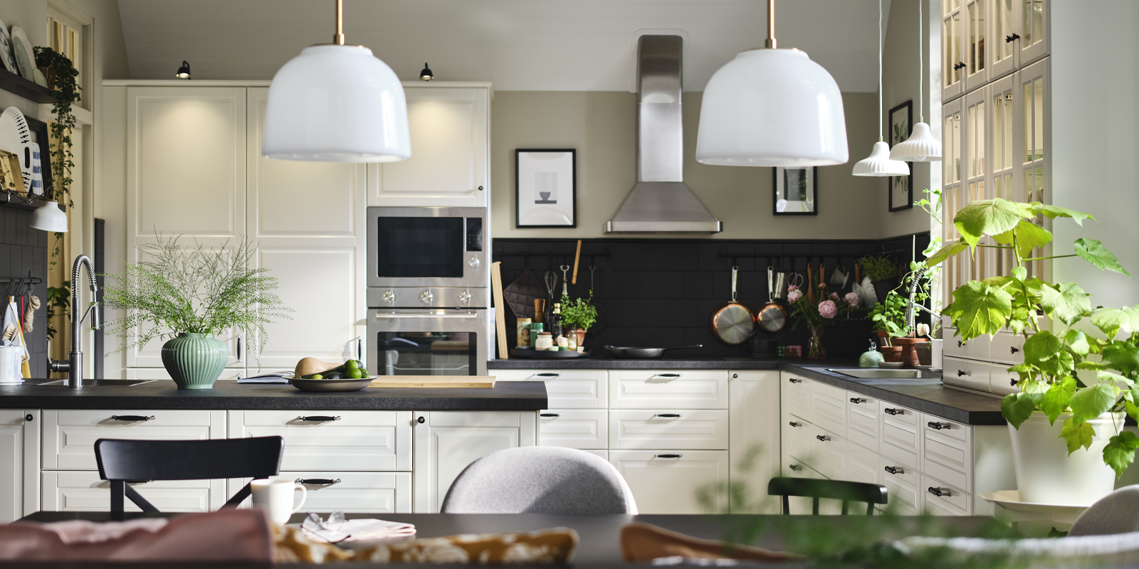 Una cocina moderna con frentes BODBYN en color blanco roto, plantas verdes y objetos decorativos, con lámparas colgantes blancas sobre una mesa.