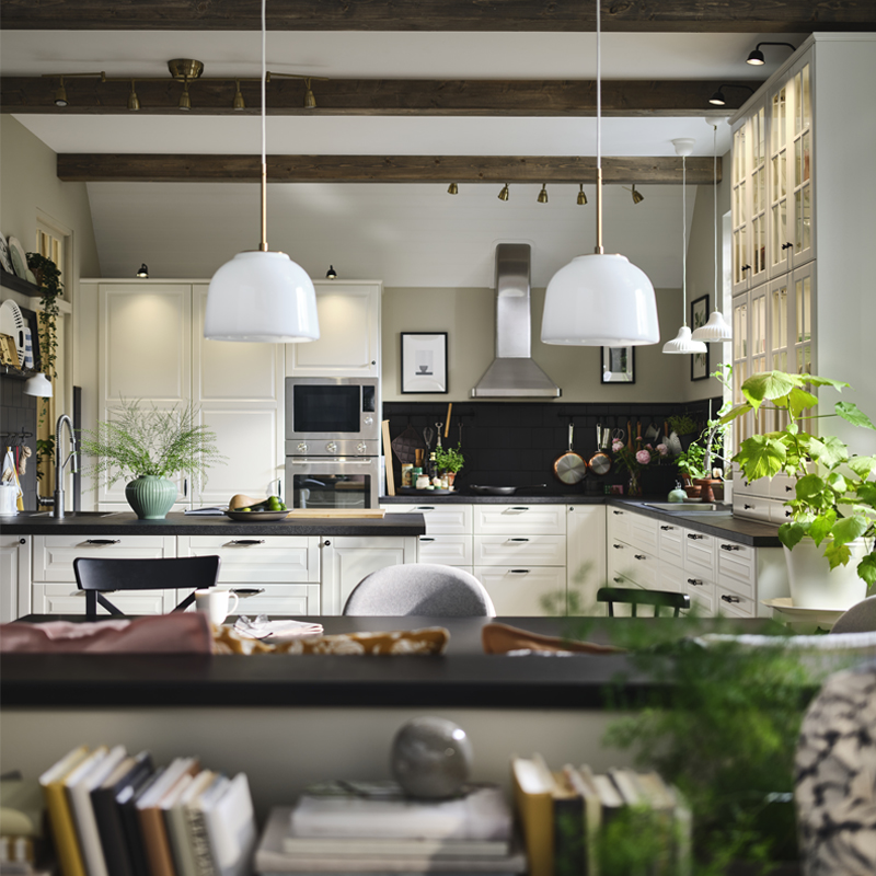 Una cocina moderna con frentes BODBYN en color blanco roto, plantas verdes y objetos decorativos, con lámparas colgantes blancas sobre una mesa.
