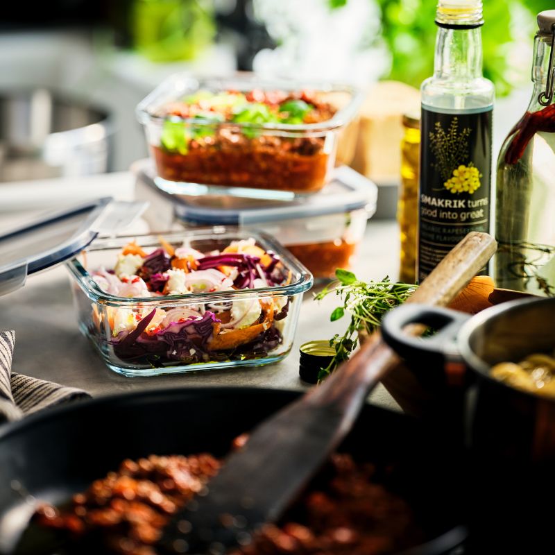 Un recipiente de vidrio IKEA 365+ para almacenar alimentos con comida preparada está junto a una sartén. Detrás de él hay otros dos recipientes apilados.