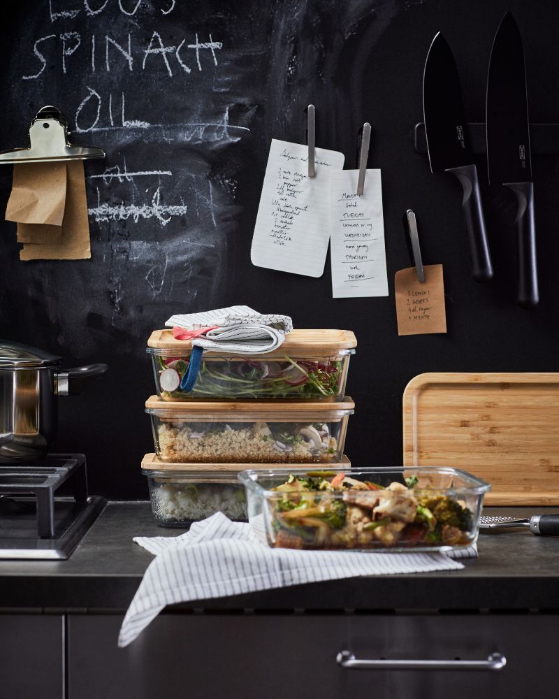 Recipientes de vidrio IKEA 365+ con tapas de bambú están sobre una encimera de cocina oscura. Detrás de los recipientes hay una pizarra negra.