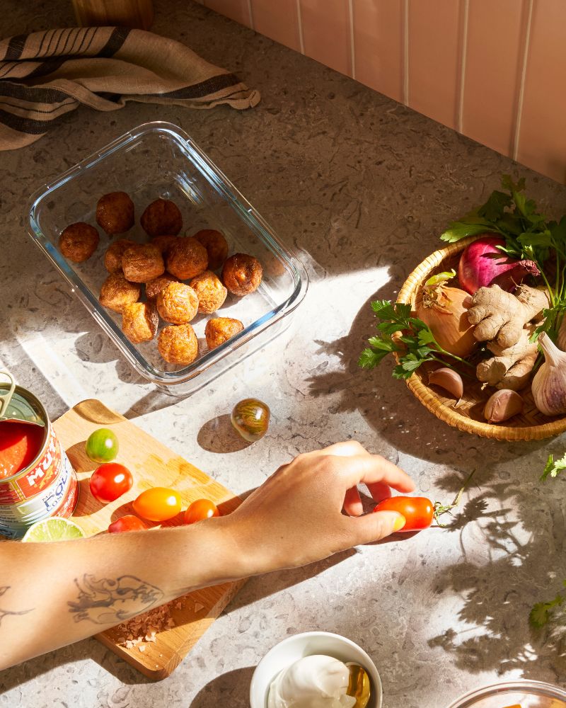 Sobre una encimera de cocina, una persona toma un tomate cherry. Cerca hay un recipiente de vidrio IKEA 365+ con albóndigas vegetales.