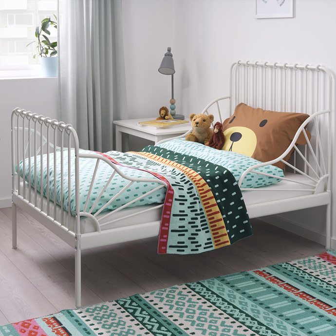 White metal children's bed with patterned bedding, bear-shaped pillow, and soft toys in a bright room.