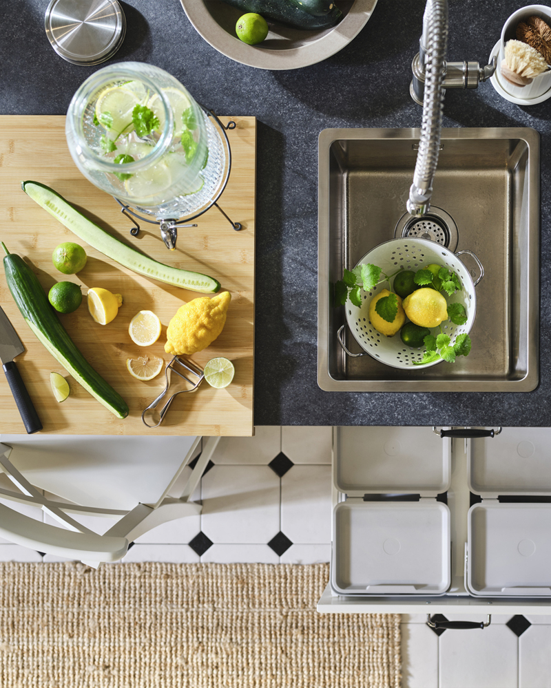 Una encimera de cocina negra con fregadero empotrado VRESJÖN de acero inoxidable, con un colador lleno de limones y hierbas.