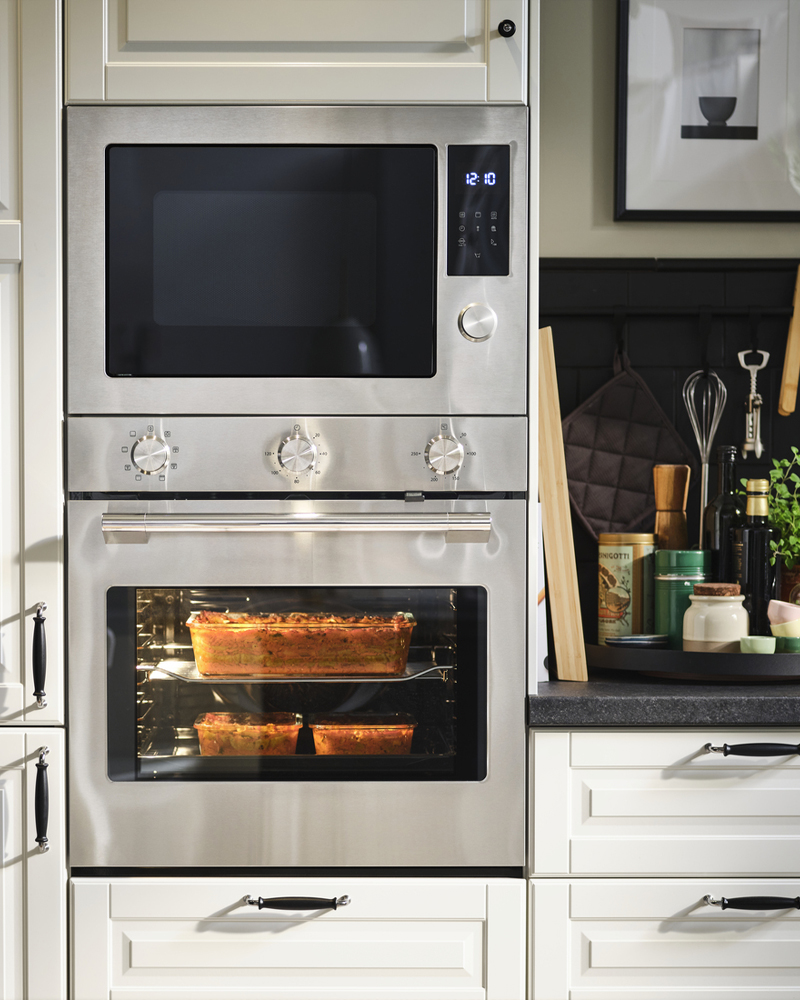 Un horno con ventilador y un microondas empotrado STENABY, ambos de acero inoxidable, se colocan en una cocina con frentes BODBYN en color blanco roto.