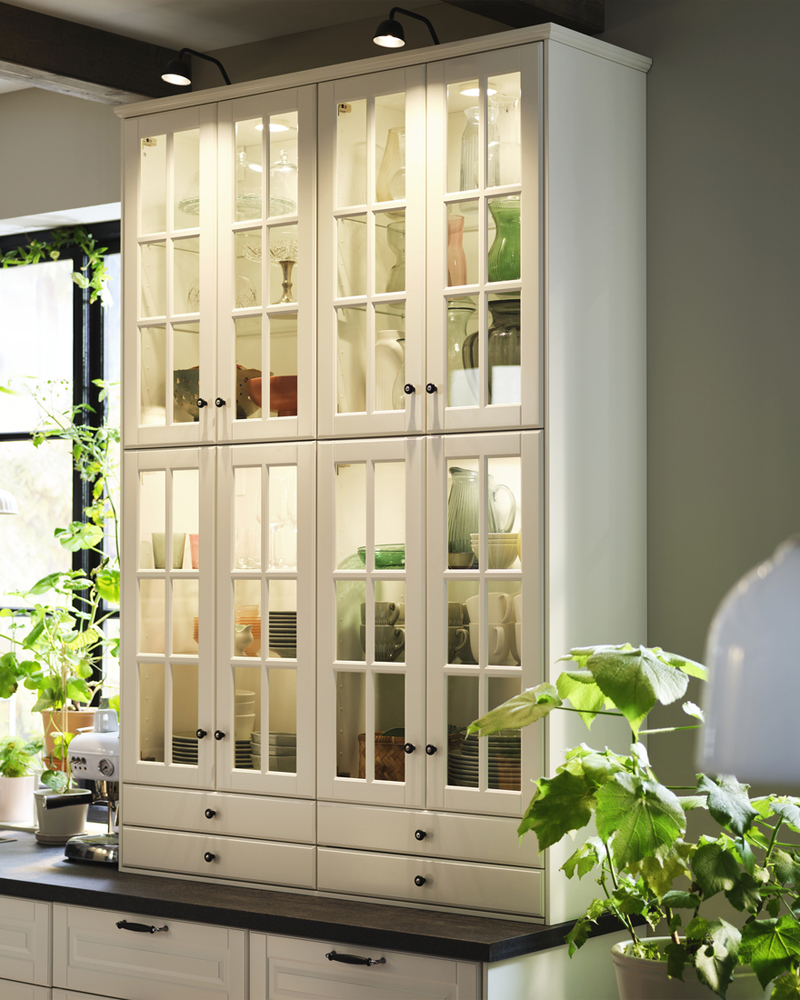 Un armario alto con puertas de cristal BODBYN en color blanco roto muestra los utensilios de cocina, rodeado de plantas en una habitación luminosa.