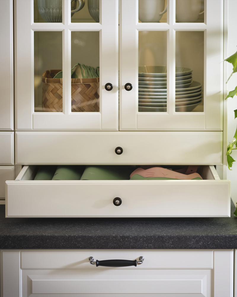 Un armario con puertas y frentes de cajones de cristal BODBYN en color blanco roto muestra platos y tazas, y ropa de hogar cuidadosamente doblada.