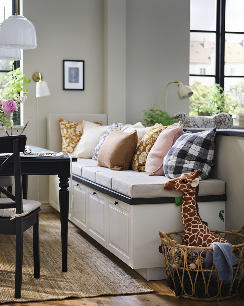 Un rincón comedor con cajones BODBYN en color blanco roto debajo de un banco, decorado con cojines, flores y una cesta con juguetes.