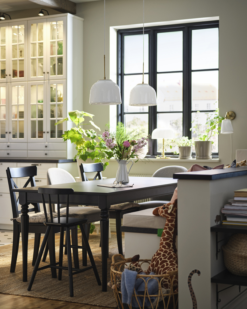 Un comedor con mesa y sillas de madera negra ROSENTORP, decorado con flores en un jarrón, junto a una cesta con peluches.