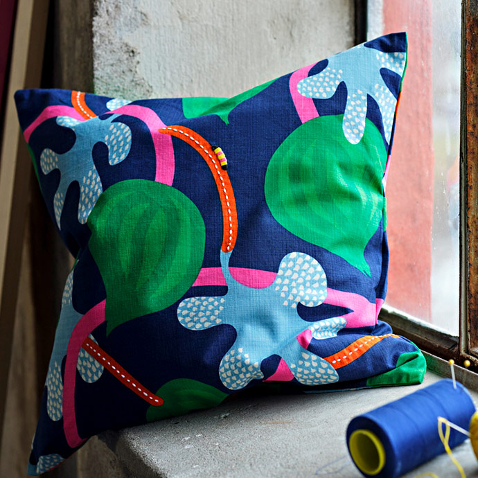 Cojín con estampado botánico en azul, verde y rosa sobre repisa de piedra junto a un carrete de hilo.