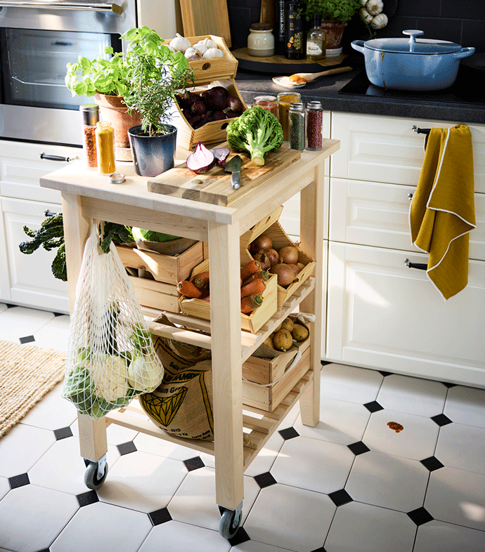 Light wood kitchen trolley on wheels organized with wooden crates of vegetables, herbs, and spices in a bright kitchen with tiled floors.