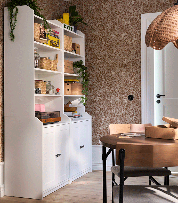 Estantería blanca con armarios inferiores organizada con cestos y libros frente a un papel pintado marrón con motivos decorativos.