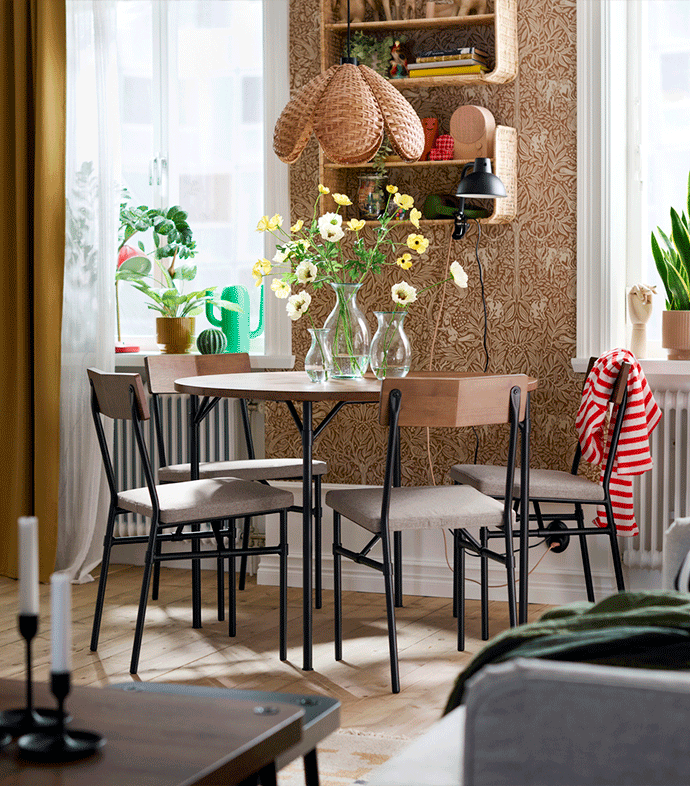 Mesa de comedor redonda de madera con cuatro sillas y una lámpara de techo trenzada en una estancia con papel pintado y luz natural.