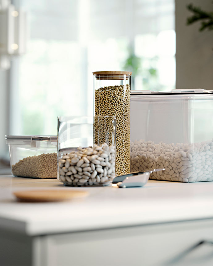 Varios botes de cristal y recipientes herméticos con granos y legumbres sobre una encimera blanca, con una pequeña pala de metal.