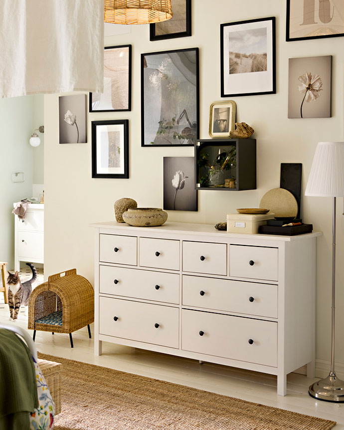 Cómoda blanca de ocho cajones en un dormitorio decorado con una galería de cuadros en la pared y una pequeña caseta de mimbre para mascota.