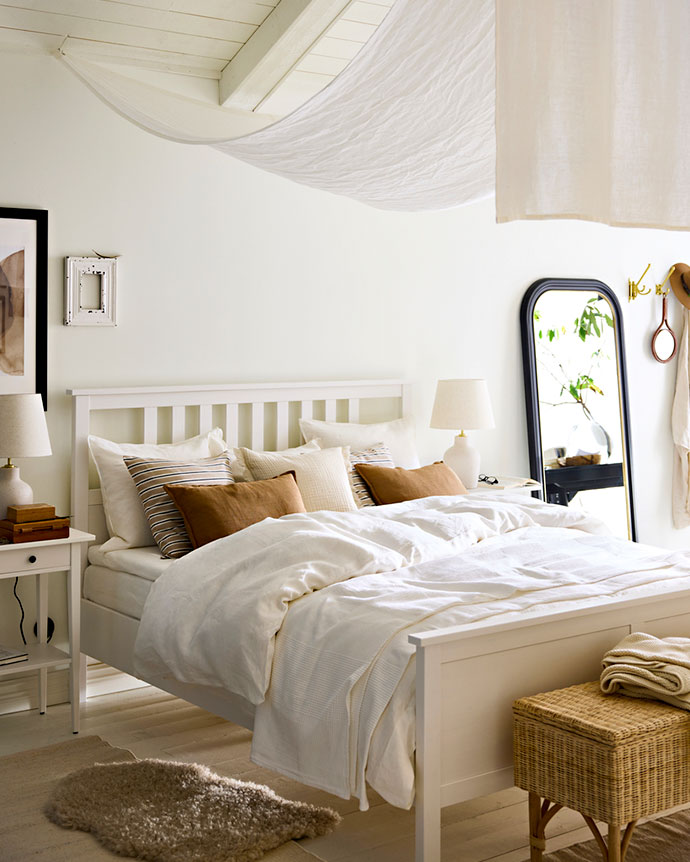 Dormitorio luminoso con cama blanca, textiles en tonos crema y tierra, un dosel ligero y un espejo de cuerpo entero con marco negro.