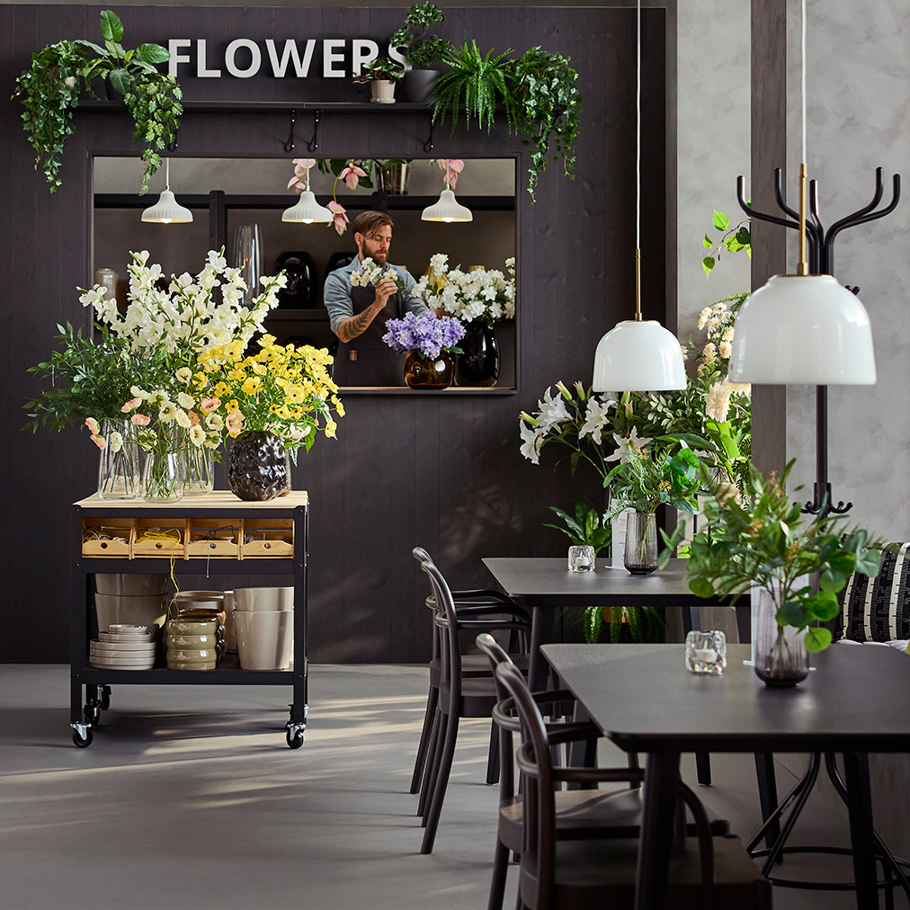 Entorno de cafetería con luz natural que incluye mesas, sillas y un carrito BROR negro/pino que exhibe flores y jarrones.