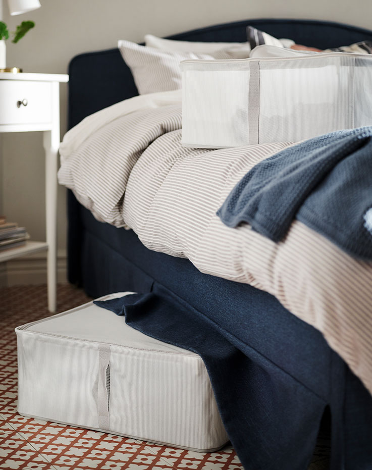 El lateral de una cama RAMNEFJÄLL con una manta y un maletín de almacenamiento HEMMAFIXARE encima. Hay otro maletín de almacenamiento debajo de la cama.