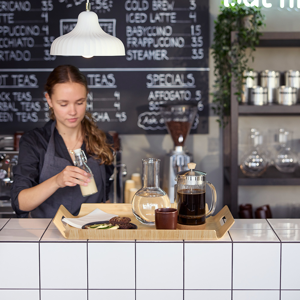 Escena de cafetería con un barista preparando una bandeja de bambú SILVERHAJMAL para servir bebidas y comida sobre un mostrador de cafetería embaldosado.