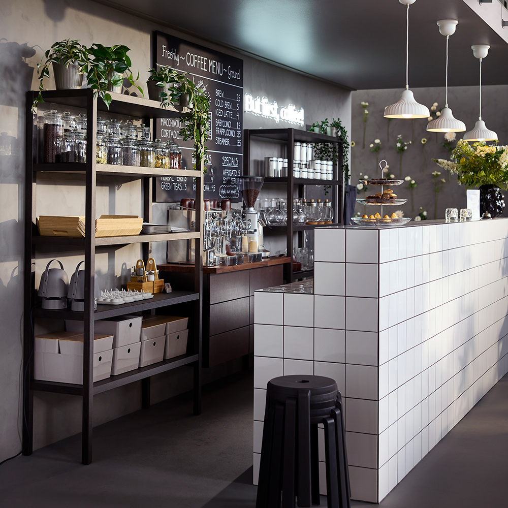 Un mostrador de cafetería con estanterías STOCKHOLM 2025 en chapa de roble/marrón oscuro, llenas de cristalería y frascos.