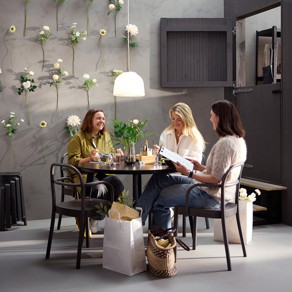 Escena de cafetería con tres personas en una mesa LISABO negra, sillas STOCKHOLM 2025 de color marrón oscuro y una pared de flores al fondo.