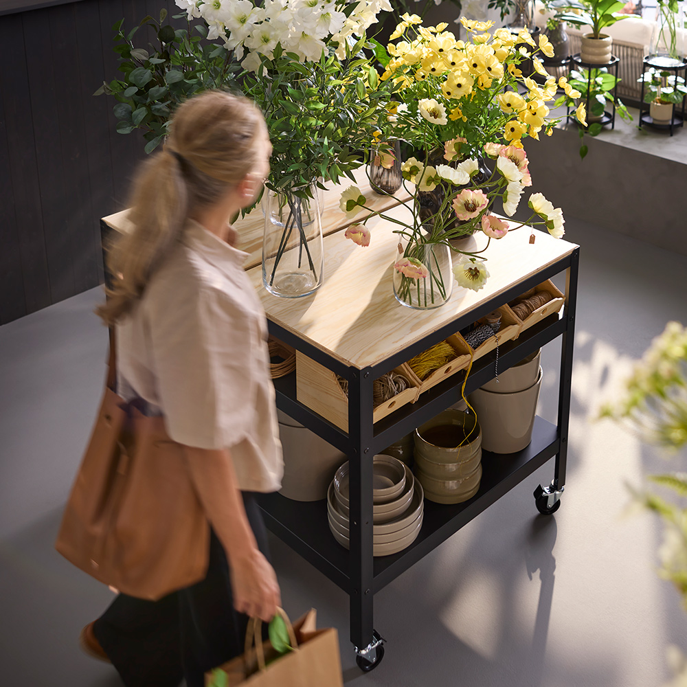 Una persona en una floristería pasando junto a un carrito BROR negro hecho de madera contrachapada de pino, que sostiene plantas y flores.