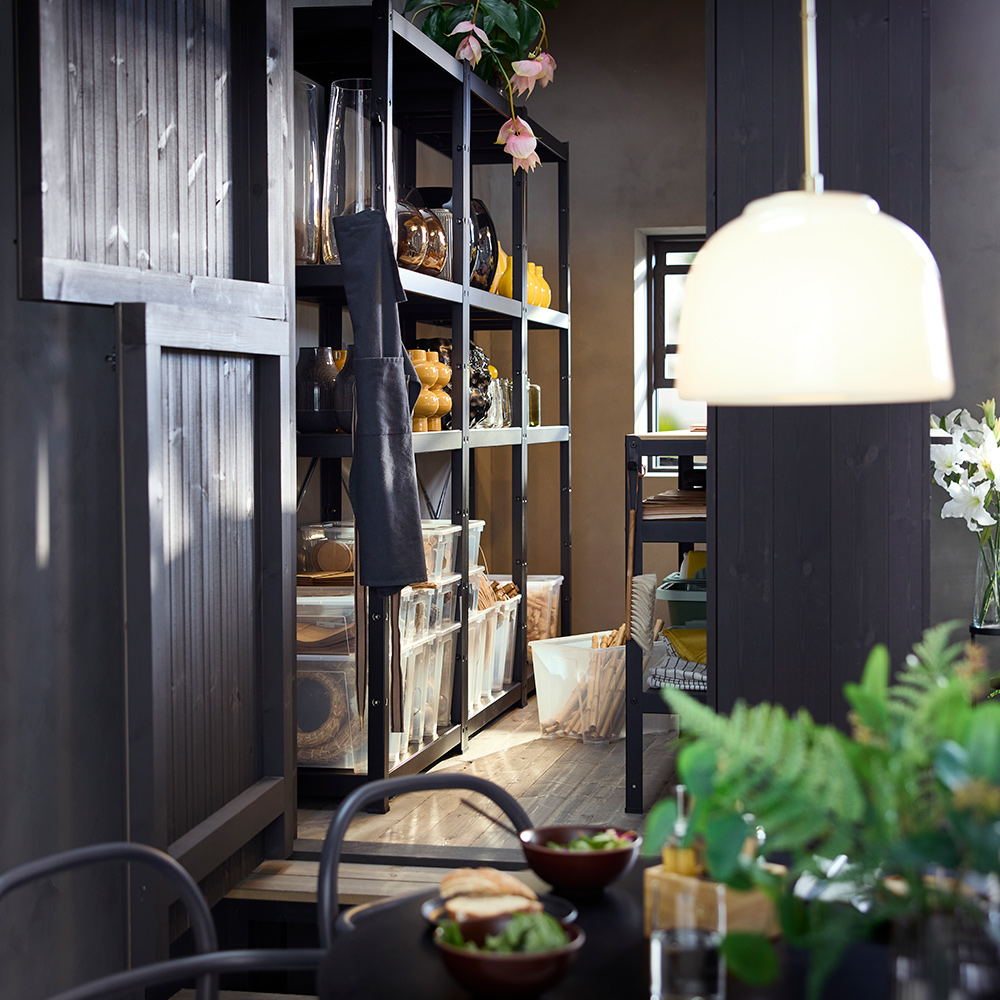 Un almacén de tienda con una estantería BROR negra que exhibe cajas transparentes, jarrones y plantas en sus estantes.