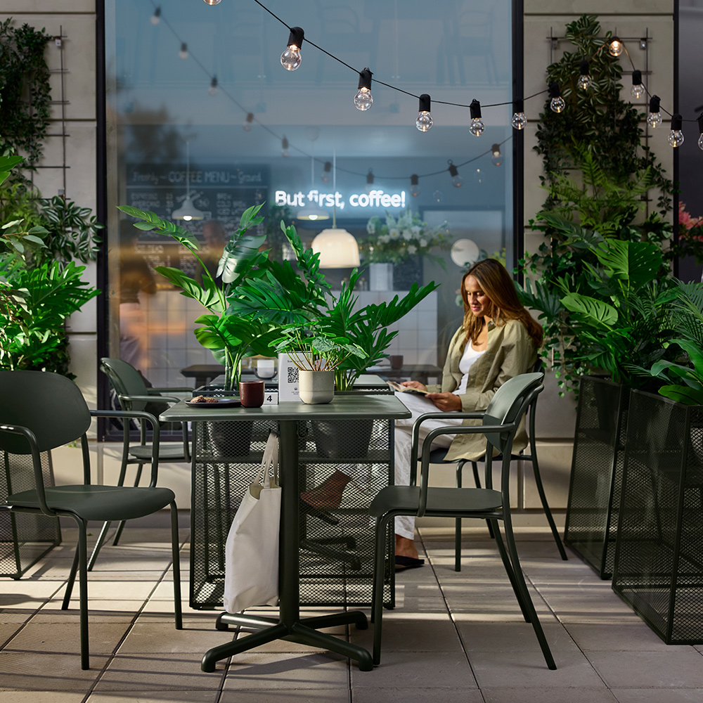 Área de servicio de cafetería al aire libre con mesas VISINGSÖ verde oscuro, sillas de metal y una persona leyendo.