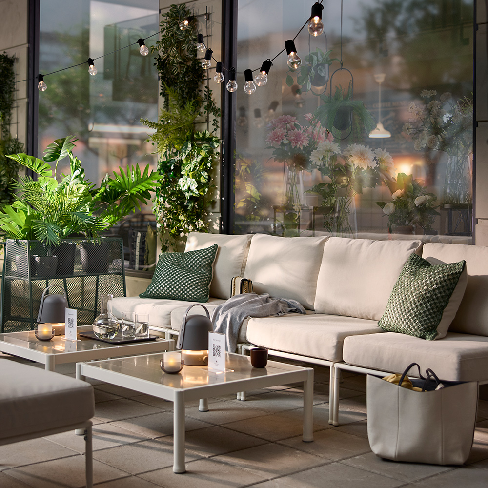 Una zona de cafetería al aire libre con un módulo de asiento SEGERÖN en blanco/beige para un sofá modular, rodeado de plantas verdes y luces acogedoras.