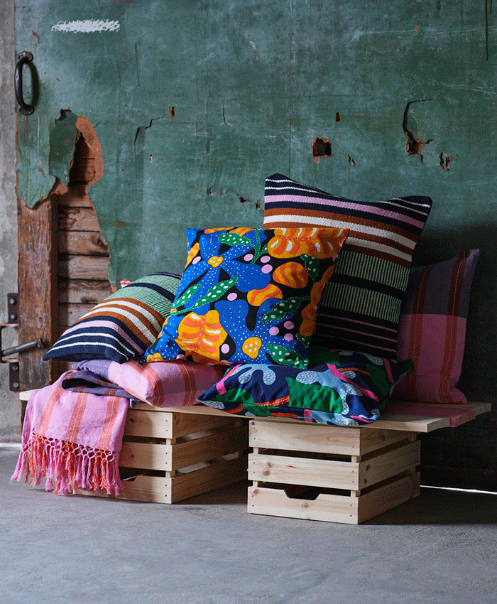 Assorted colorful cushions with floral and striped patterns stacked on wooden crates against a distressed green wall with a pink fringed throw.