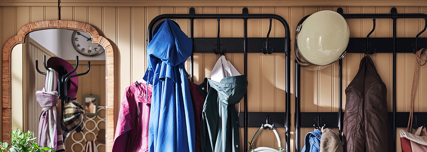 Wall-mounted black metal coat racks with hooks holding various jackets and a helmet, next to a wicker-framed mirror in a beige paneled hallway.