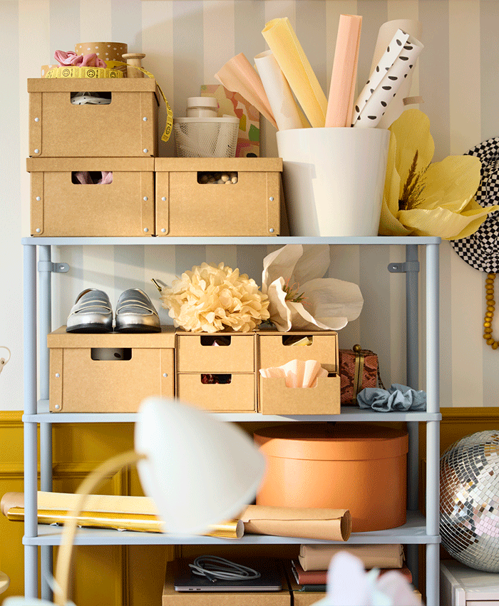 Light blue metal shelving unit organized with various brown cardboard boxes, rolls of paper, a large fabric flower, and silver shoes.