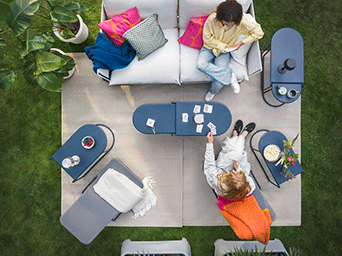Top-down view of a cozy patio with a white sofa, blue oval coffee tables, and colorful cushions on a beige rug over green grass.