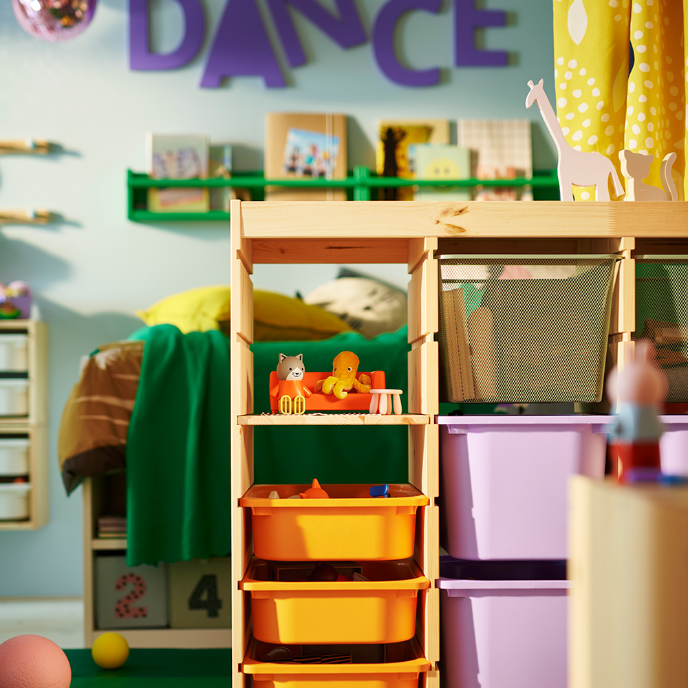 A TROFAST room divider in the middle of a children’s shared bedroom holding orange, lilac and mesh boxes, with toys on top.