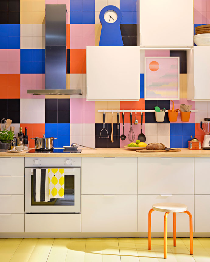 Cocina moderna con armarios blancos y pared de azulejos de colores vibrantes. Incluye encimera de madera y taburete naranja.