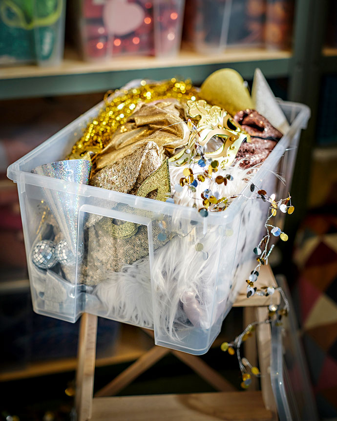Caja de plástico transparente llena de artículos de fiesta dorados y plateados, como máscaras y guirnaldas, sobre una escalera.