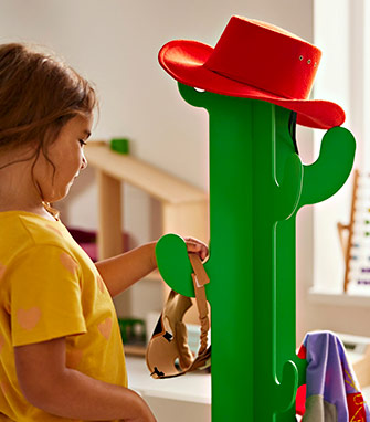 Perchero infantil verde con forma de cactus, decorado con un sombrero rojo, mientras un niño interactúa con él en una habitación soleada.