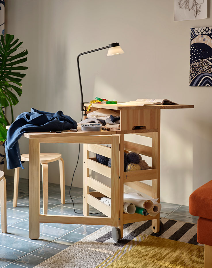 A sunlit living room with a pine RESARÖ mobile drop leaf table, leaves out and a project involving textiles placed on top.