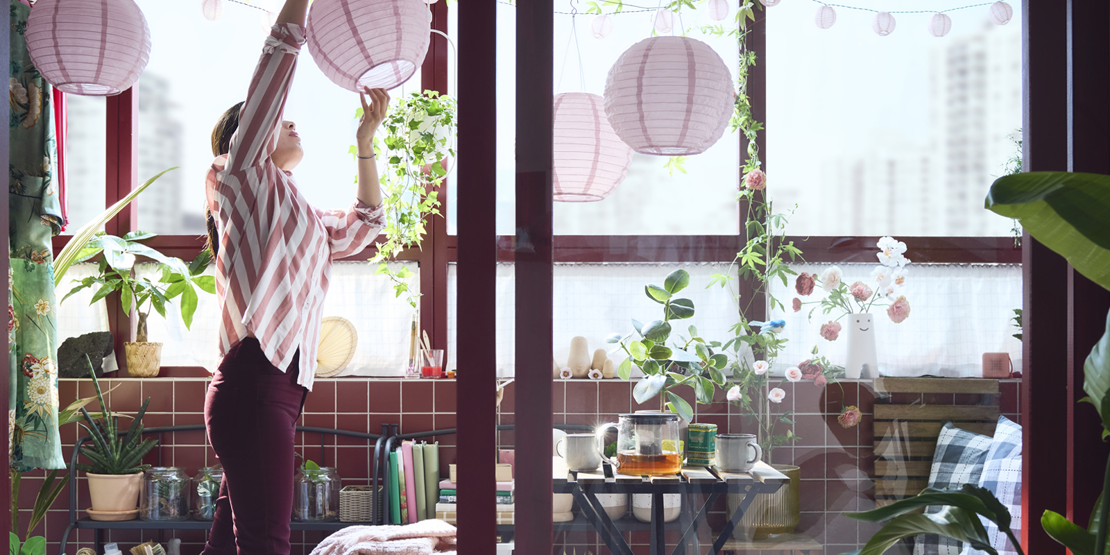 An enclosed balcony with a black and light brown TÄRNÖ outdoor table, surrounded by plants and decorative pink lamps.