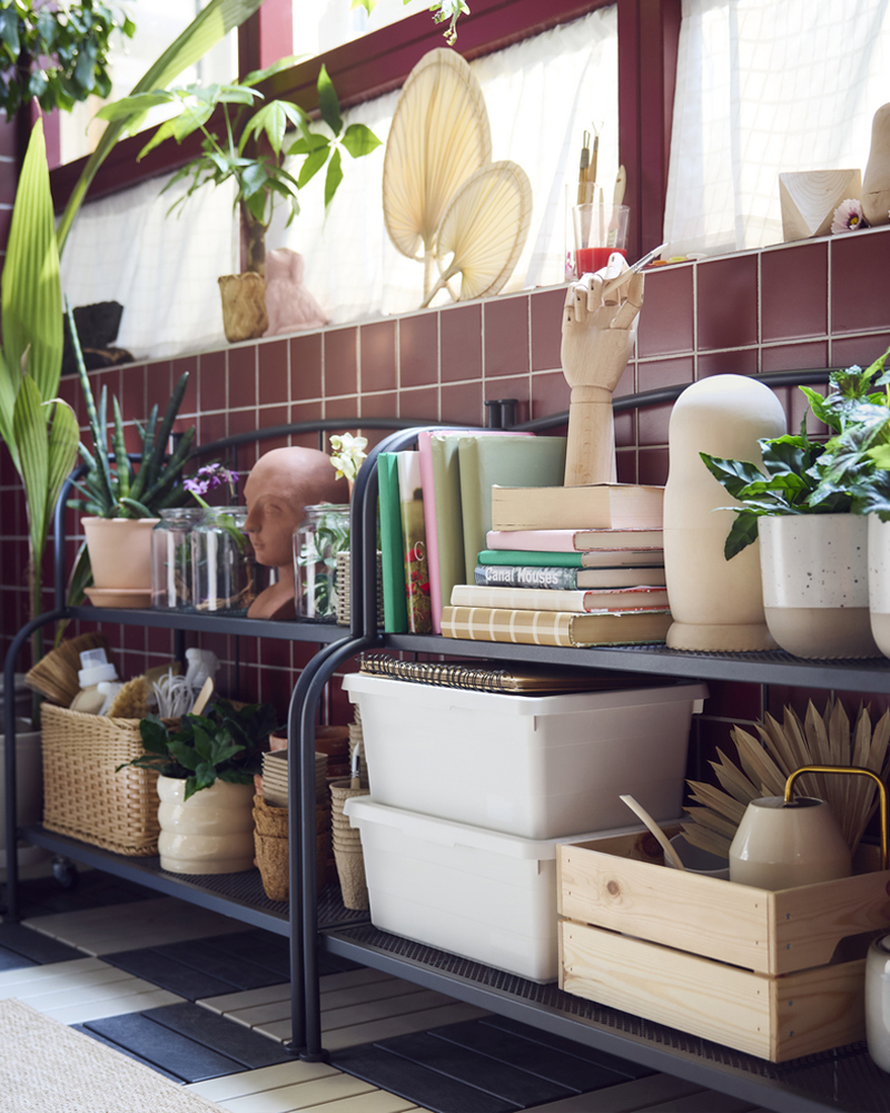 A bright enclosed balcony with two grey LÄCKÖ shelving units made for outdoor use, displaying various plants and books.